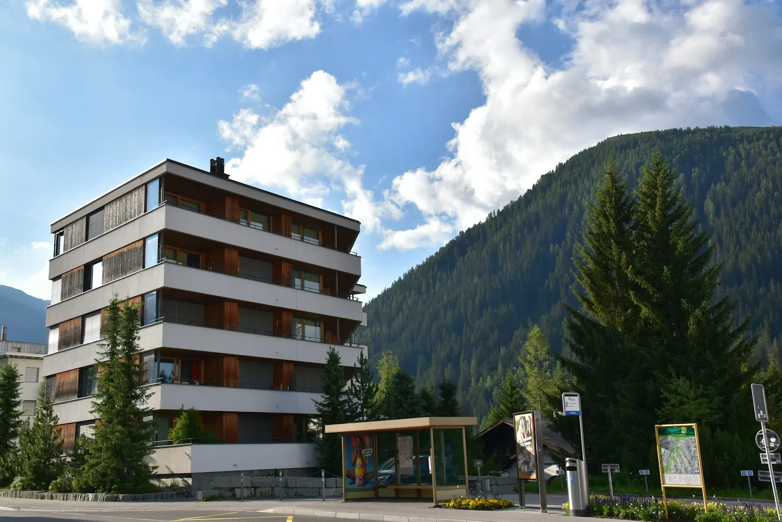 Ein modernes Mehrfamilienhaus mit umlaufenden Balkonen vor einem bewaldeten Berghang.