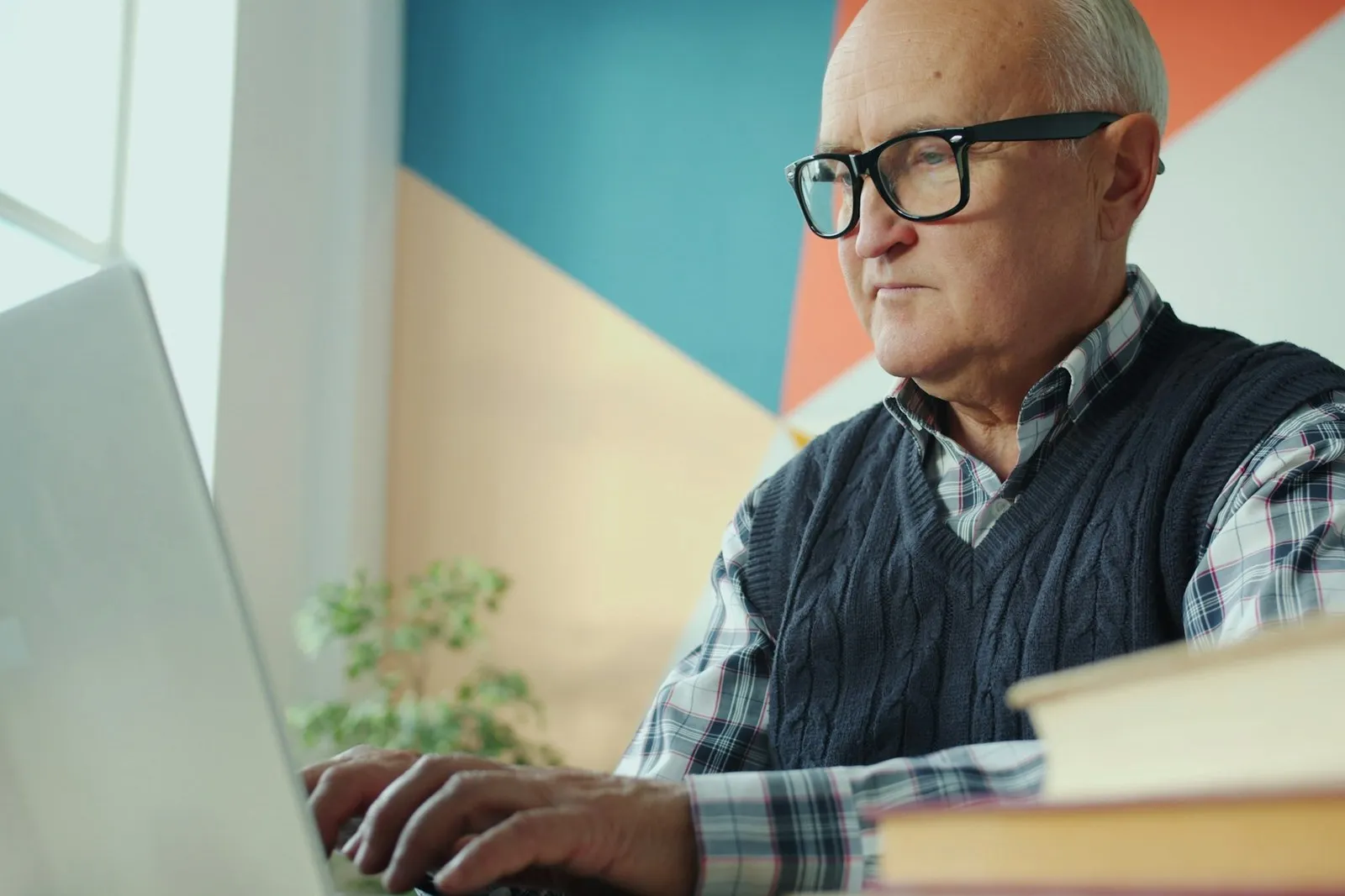 Ein älterer Mann mit Brille arbeitet konzentriert an einem Laptop, neben ihm ein Stapel Unterlagen.